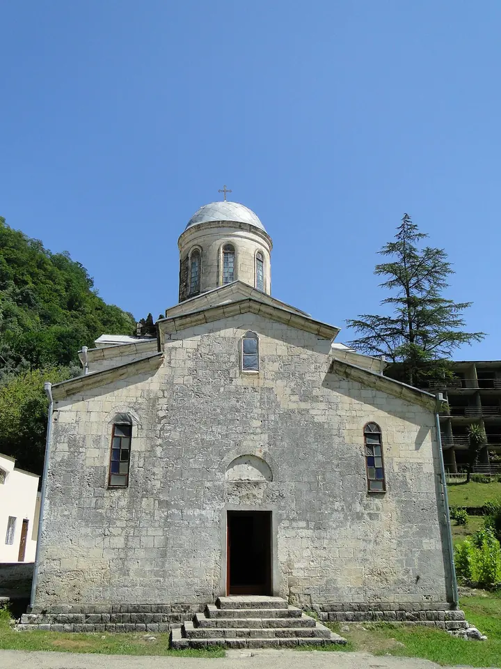 A small medieval stone church with a round drum and dome under a blue sky, surrounded by trees on a hillside