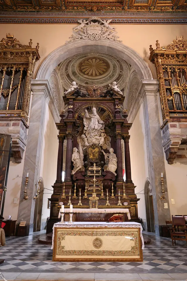 Tall Baroque high altar of dark marble and gilt bronze with a white marble statue of a pope at the top, seen down a nave with a checkered floor