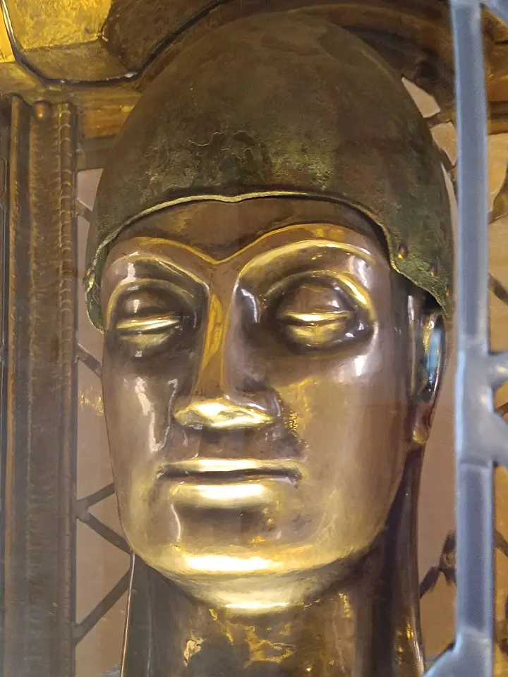 Close view of a polished gold head reliquary with a calm, smooth face and closed eyes, behind a protective grille