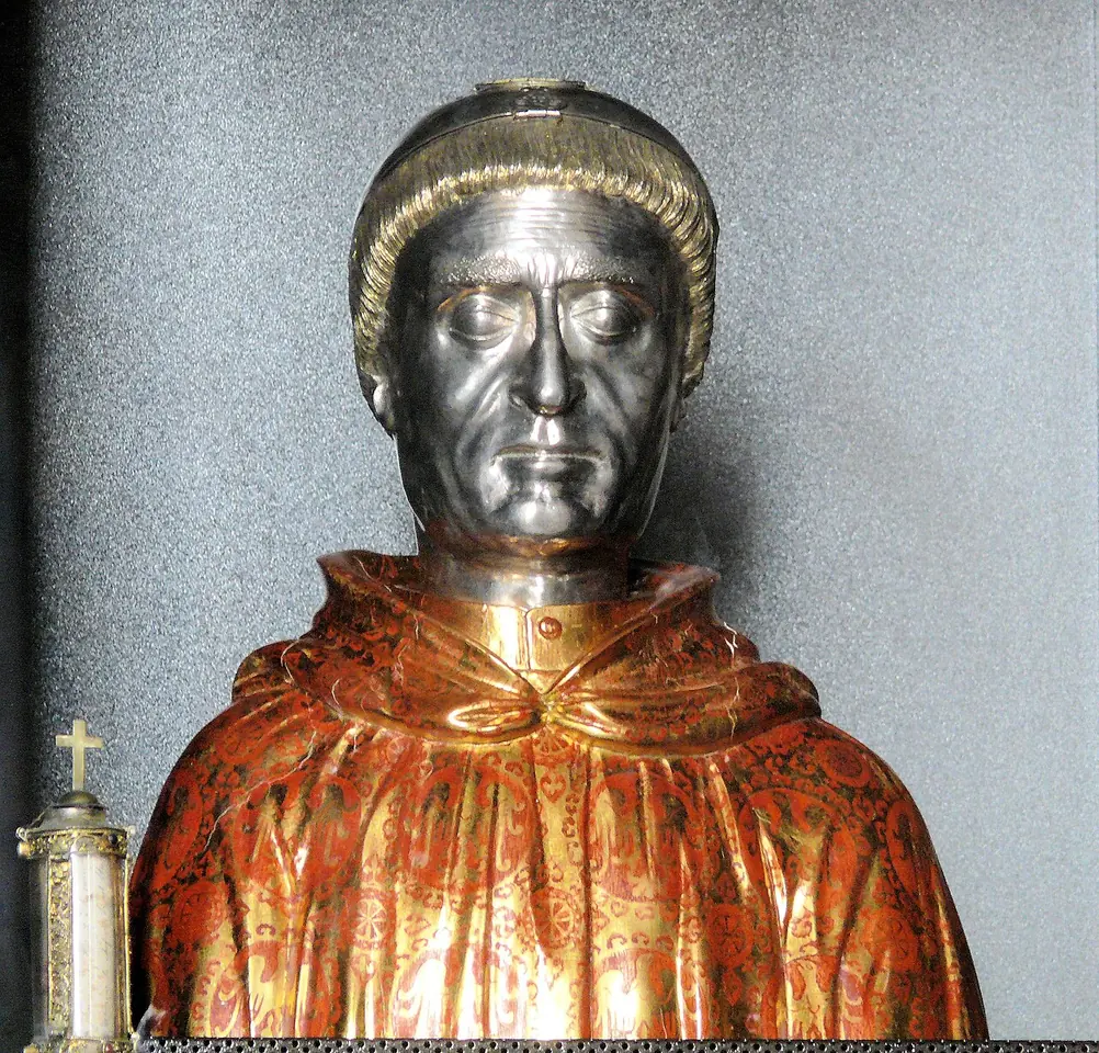 Silver reliquary bust of a tonsured man with a stern face, wearing a gilded red and gold cope, beside a small reliquary cross