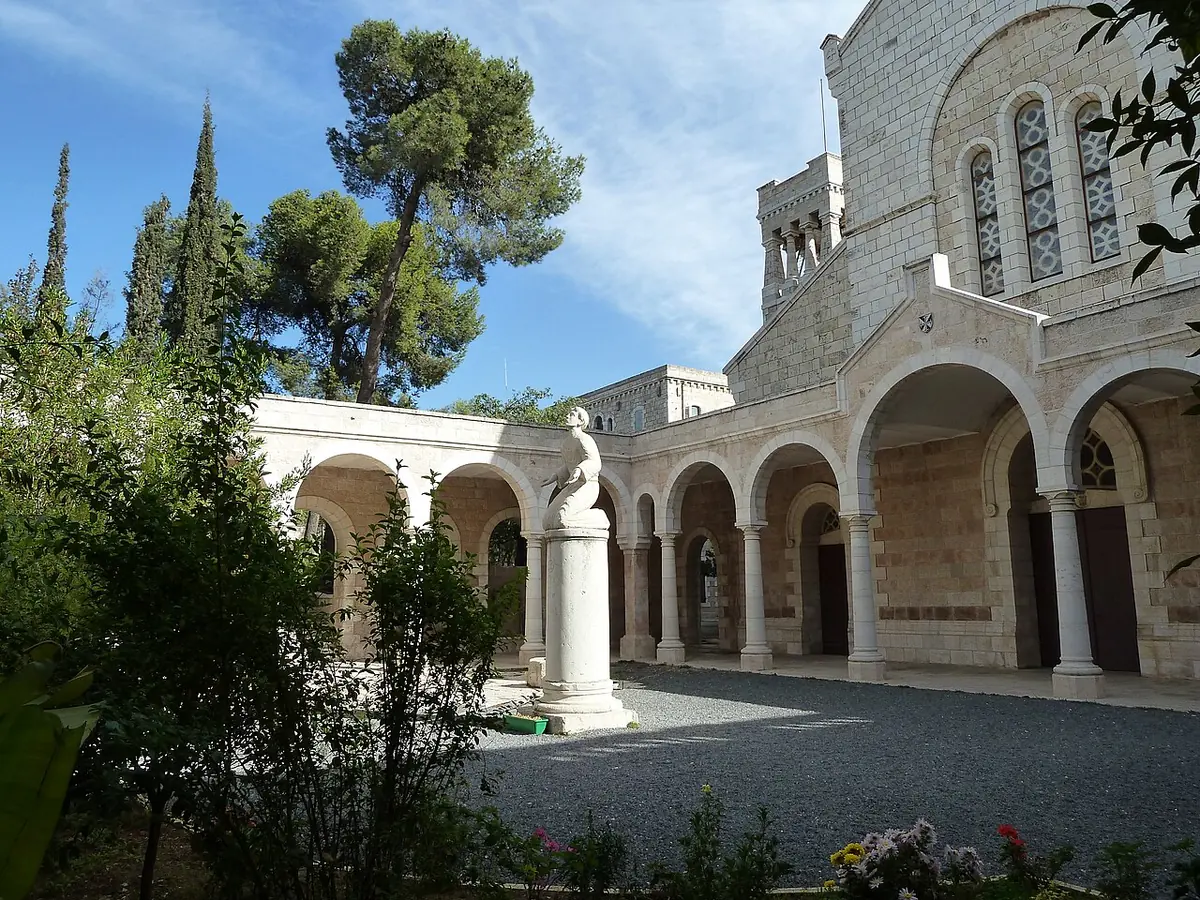 The courtyard of the Basilica of Saint-Étienne in Jerusalem, with a white statue of St. Stephen, stone arcades, cypress and pine trees