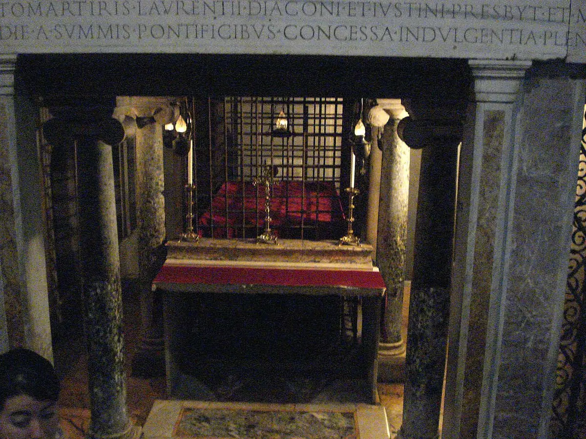 A small crypt chapel under a high altar, with a grated opening between marble columns lit by candles, and a Latin inscription above naming the martyr Lawrence