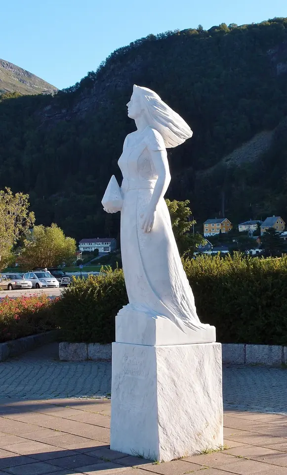 Modern white marble statue of a slender young woman with flowing hair, holding a stone in her hands, standing by a harbor with green mountains behind