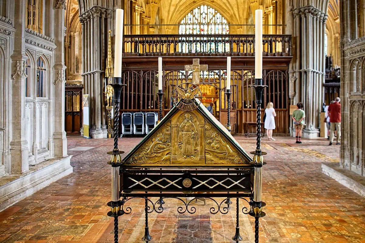 A gilded, gabled metal shrine canopy on a wrought-iron stand surrounded by tall candlesticks, on the medieval tiled floor of a Gothic cathedral retrochoir
