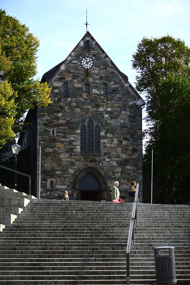 The gray stone west front of a Romanesque cathedral with a pointed gable, clock and arched door, at the top of a broad flight of steps between trees