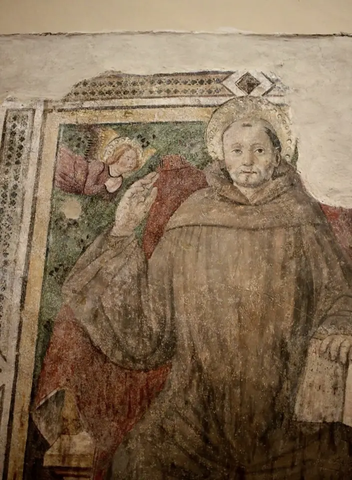 Faded fresco of a haloed elderly monk in a brown habit with his hand raised, an angel at his shoulder, framed by a painted border