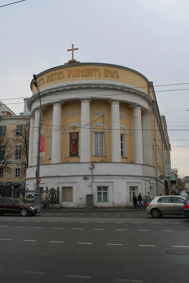 A pale yellow rounded corner building with white columns, topped by a cross, on a Moscow street
