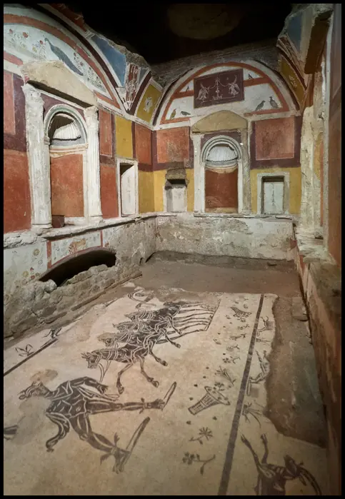 Interior of an ancient Roman tomb chamber with painted red and yellow walls, arched niches and a mosaic floor, a few meters from St. Peter's grave