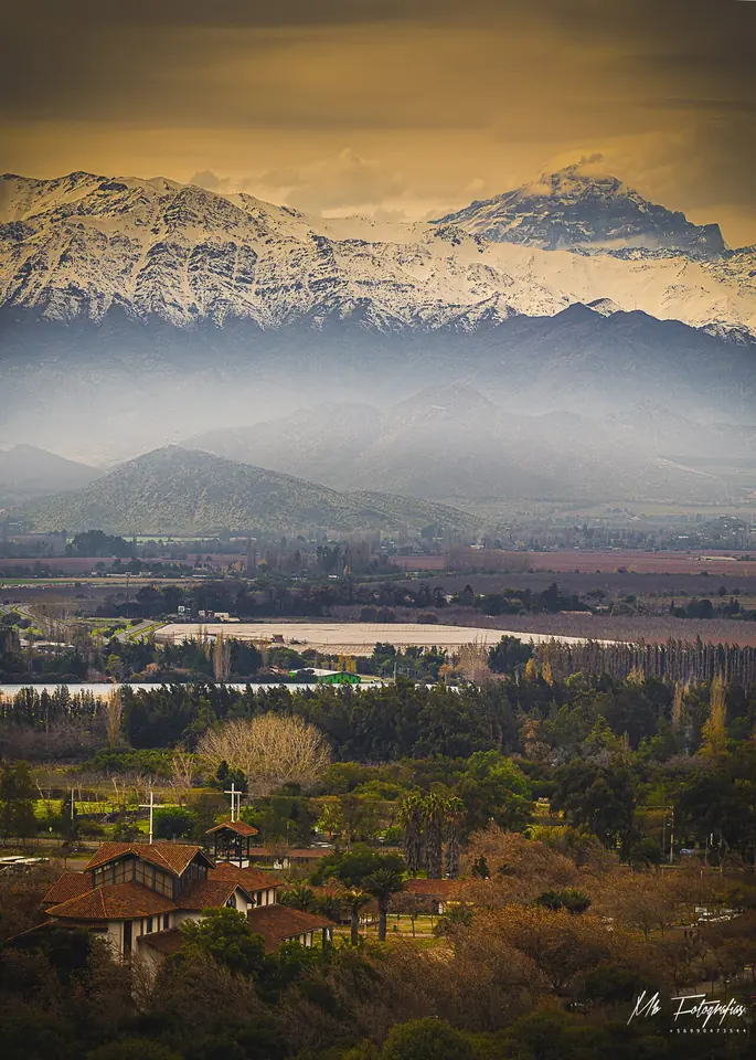 The tiled roofs of the shrine at Auco among trees in a green valley, with the snow-covered Andes and Mount Aconcagua rising behind