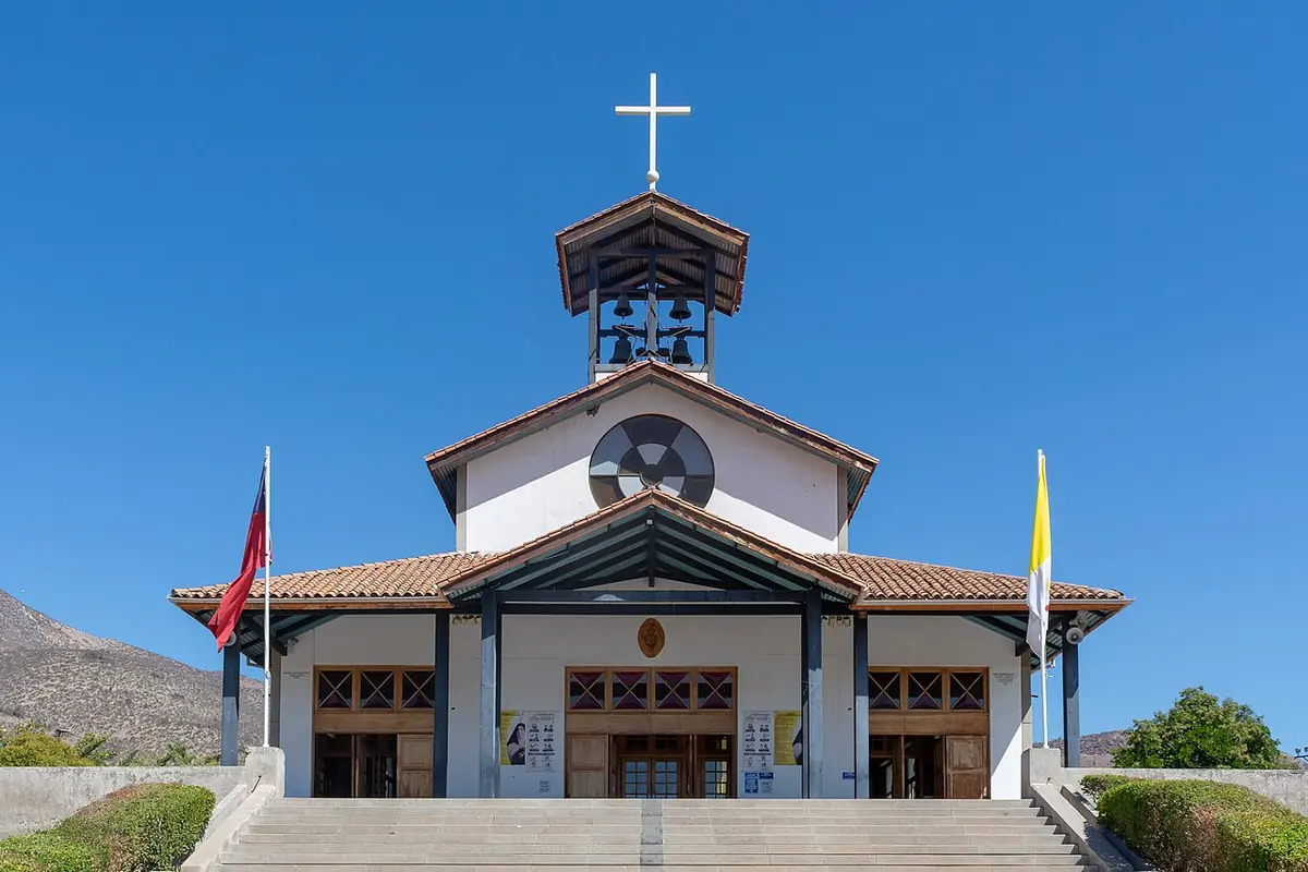 White facade of the shrine church at Auco with a bell tower and cross, a round window and wide steps, under a clear blue sky