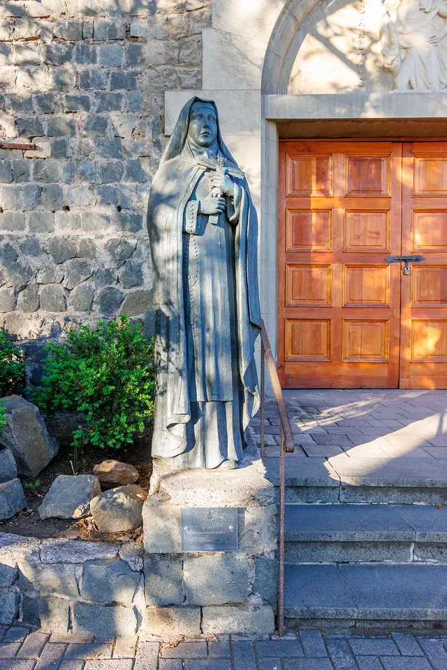 A dark metal statue of St. Teresa of Los Andes in a Carmelite habit and veil, holding a crucifix, beside a stone wall and a wooden church door