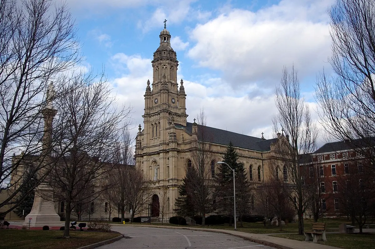 A large pale stone church with a tall domed bell tower and a long nave, seen across a curving road among bare trees