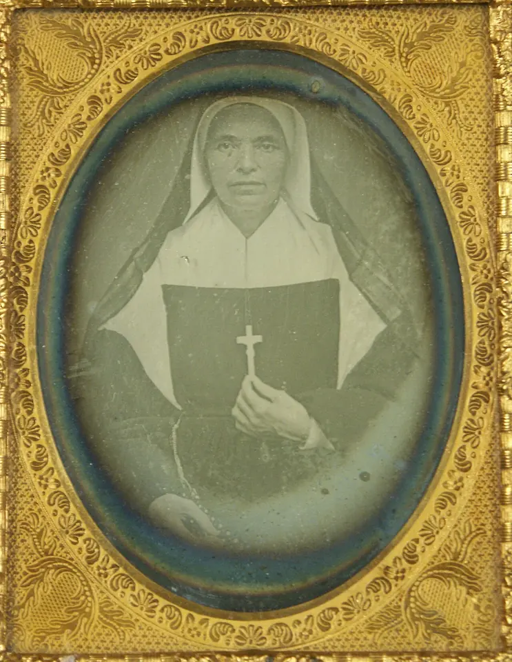 Daguerreotype in a gilt case of a sister in a dark habit and white bonnet, holding a crucifix against a black book
