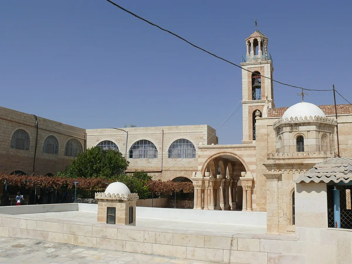 Pale stone courtyard of a monastery with arched cloisters, a small domed shrine and a tall bell tower under a clear blue sky