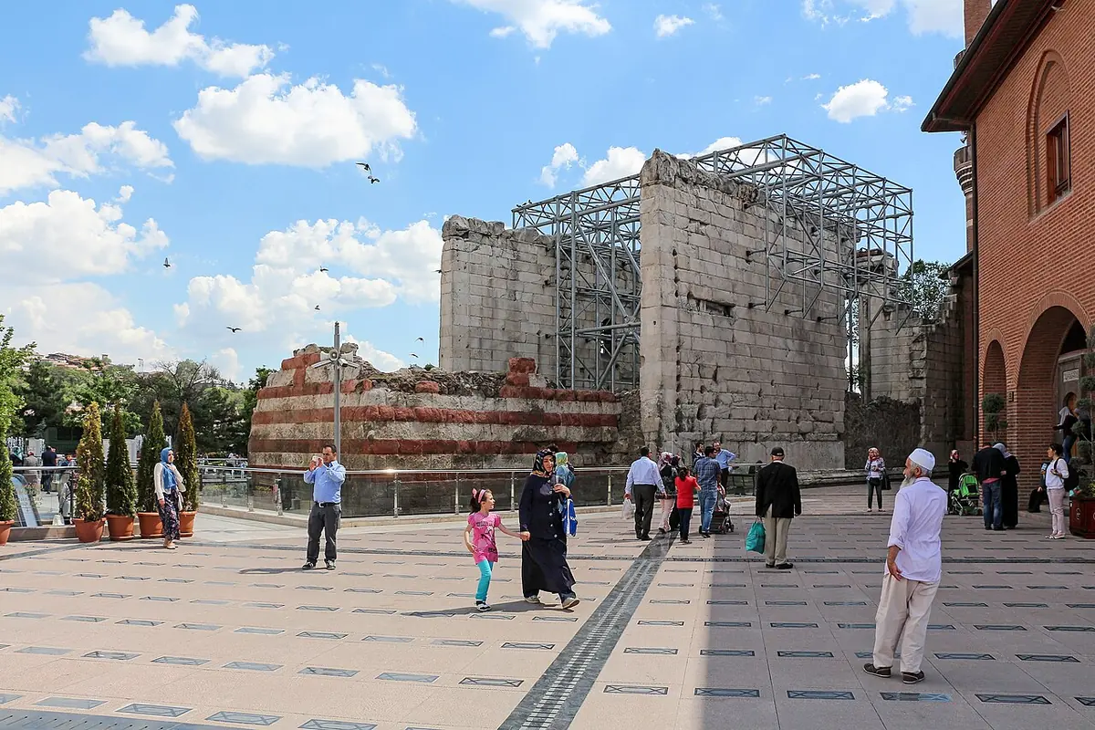 Tall weathered marble walls of a Roman temple under a metal canopy, beside a city square with people walking in the sun