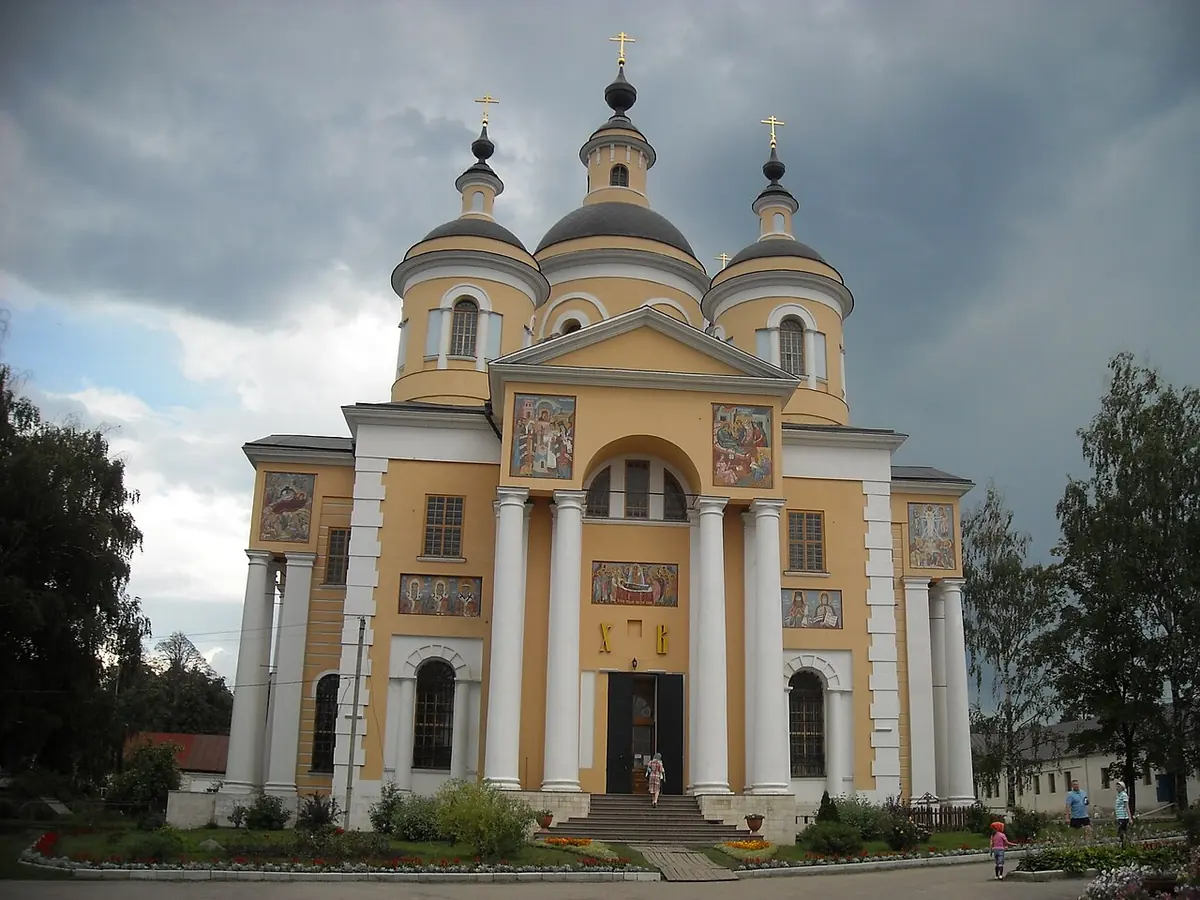 A large yellow cathedral with white columns, a portico and several dark domes topped with crosses, surrounded by trees