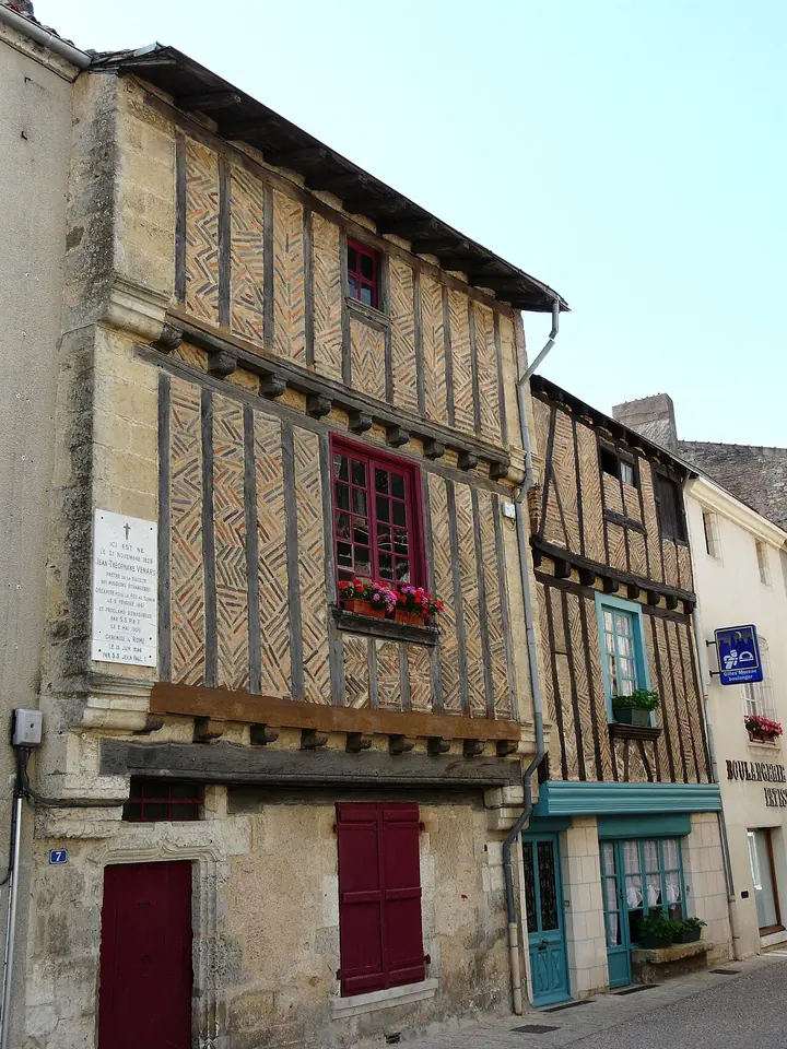 A tall old half-timbered house with a slate roof and red shutters on a narrow village street, a white plaque on its wall