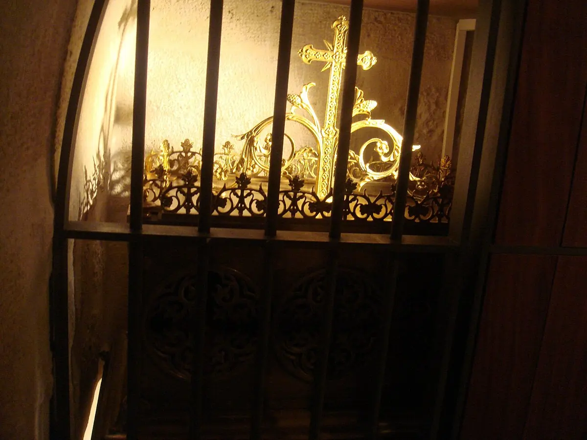 A dim crypt niche behind dark iron bars, lit from within, with a gilded cross and scrollwork above a reliquary chest