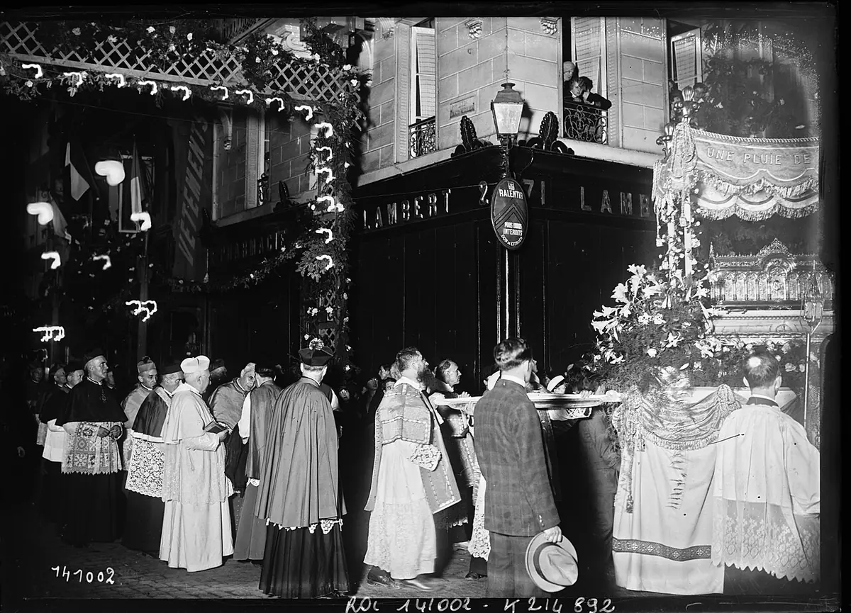 Black-and-white press photograph of priests in vestments beside St. Thérèse's flower-decked reliquary on a lit, decorated street corner in Lisieux in 1929