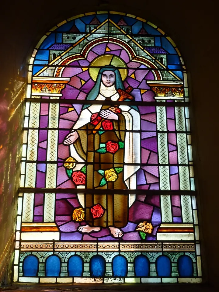 Stained-glass window of St. Thérèse in her Carmelite habit against a purple background, holding red roses with more roses falling at her feet