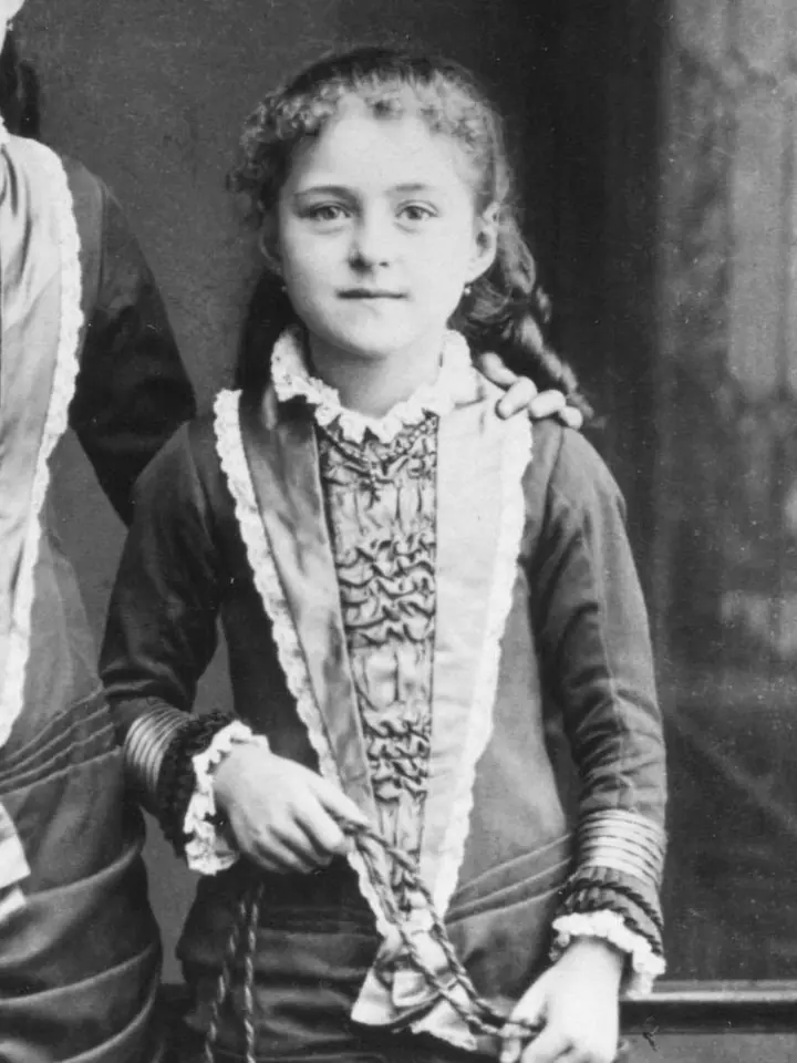 Fotografía en blanco y negro de estudio de una niña de ocho años de cabello rizado, con un vestido de volantes y una cuerda de saltar en la mano