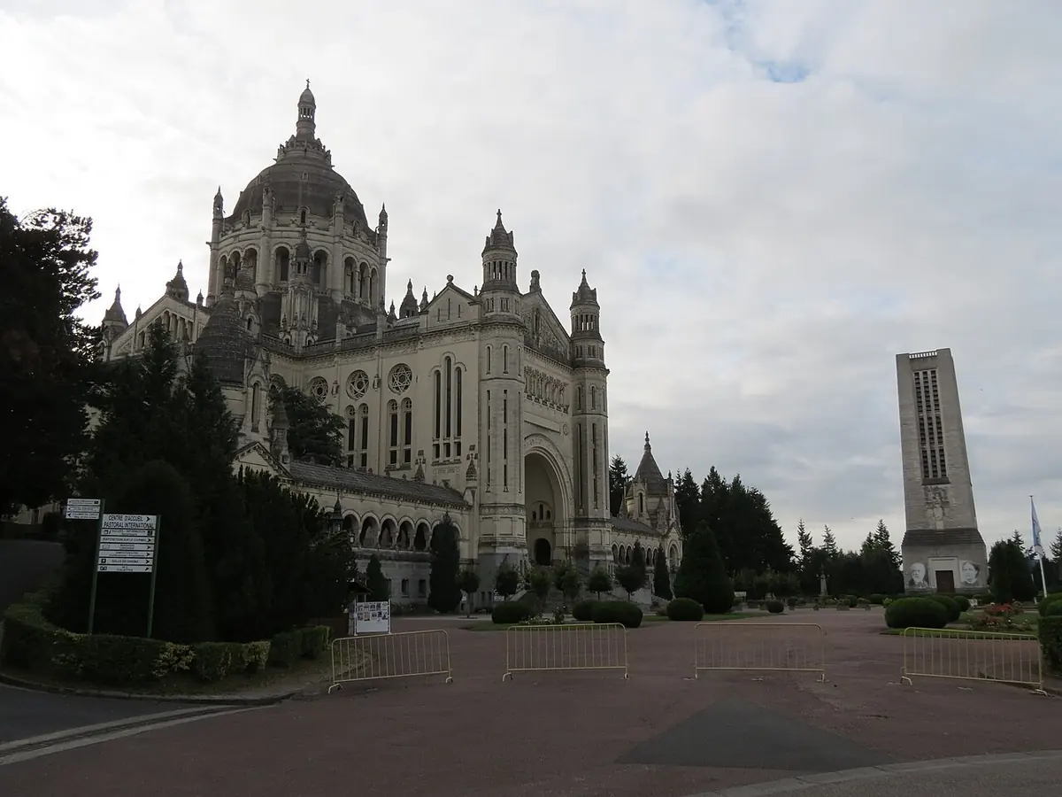 Gran basílica de piedra clara con cúpula y un alto campanario cuadrado al lado, vista desde una amplia explanada