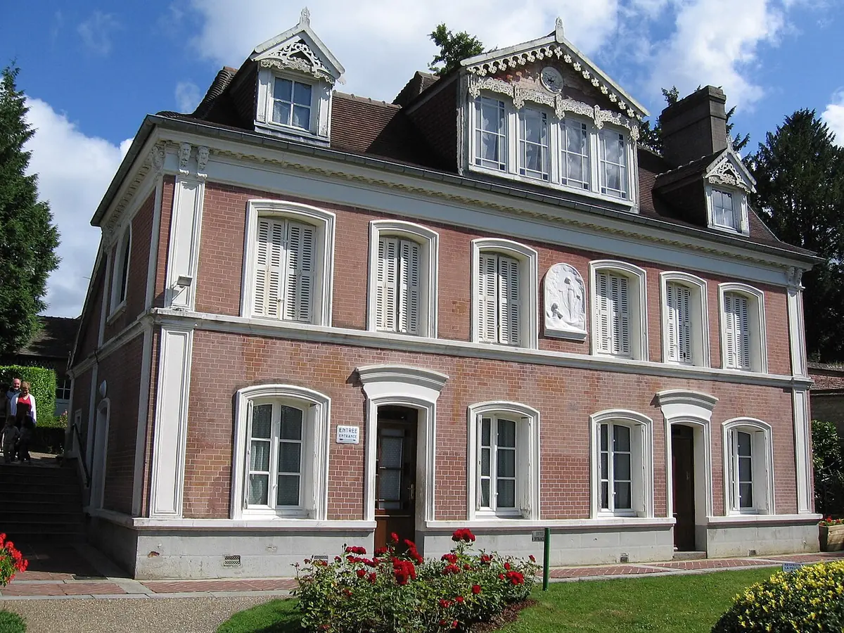 Casa de ladrillo de dos pisos con ventanas de marco blanco, techo con buhardillas y un macizo de flores delante
