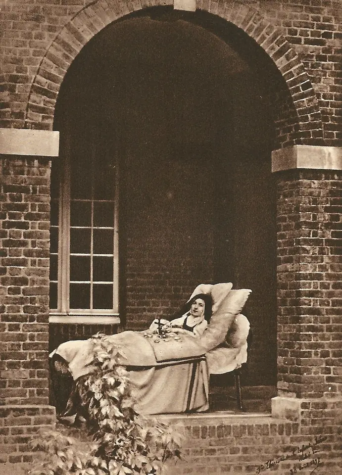 Fotografía sepia de una joven monja acostada en una cama bajo un arco de ladrillo del claustro, con un crucifijo en las manos y pétalos de rosa sobre la colcha