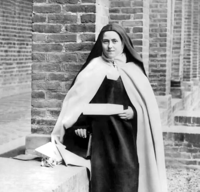 Fotografía en blanco y negro de una joven carmelita con capa blanca y velo negro, sentada en un escalón junto a unos arcos de ladrillo, con un rollo escrito y un libro en las manos