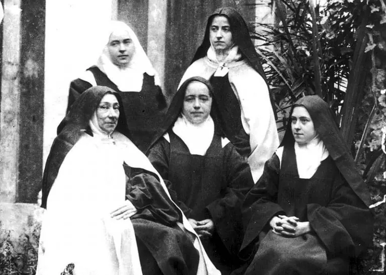 Fotografía en blanco y negro de cinco monjas carmelitas con hábito y velo, tres sentadas y dos de pie, en el patio de un jardín