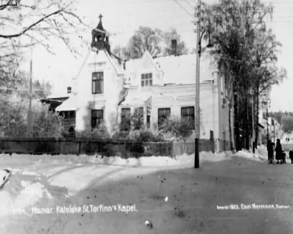 Old black-and-white photograph of a white wooden villa with a small bell turret, set behind a fence on a snowy street