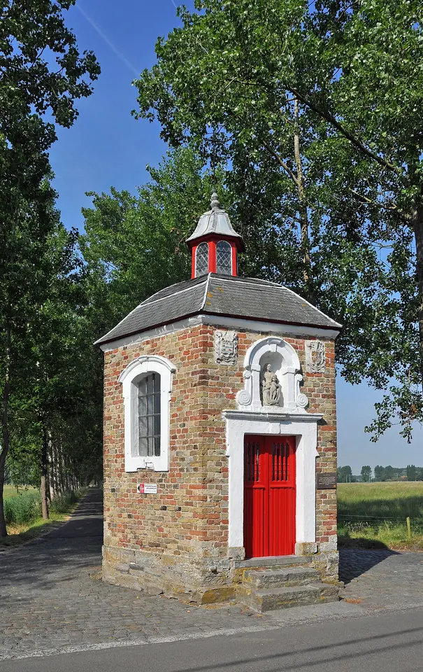 A small square brick wayside chapel with a red door, a white-framed window and a little lantern on its roof, beside a country road