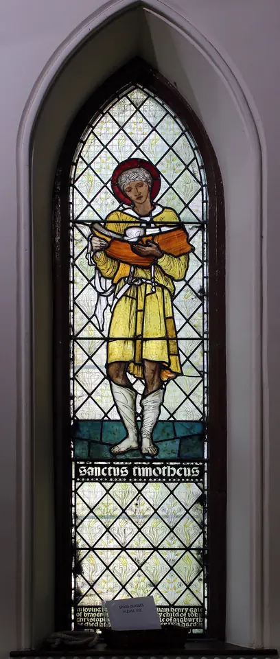 Narrow pointed stained-glass window of a young St. Timothy in a yellow tunic and halo holding an open book, with the words Sanctus Timotheus below