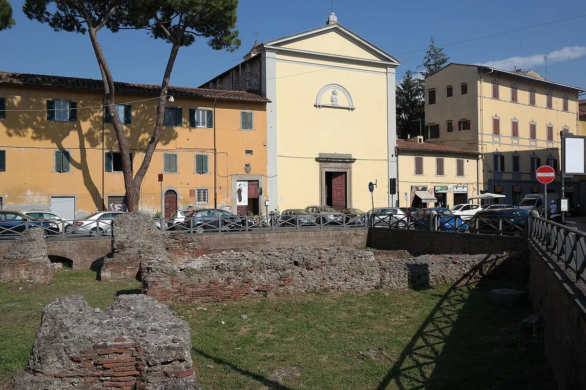 Low brick ruins of Roman baths in a grassy enclosure under pine trees, with a plain pale yellow church facade behind them