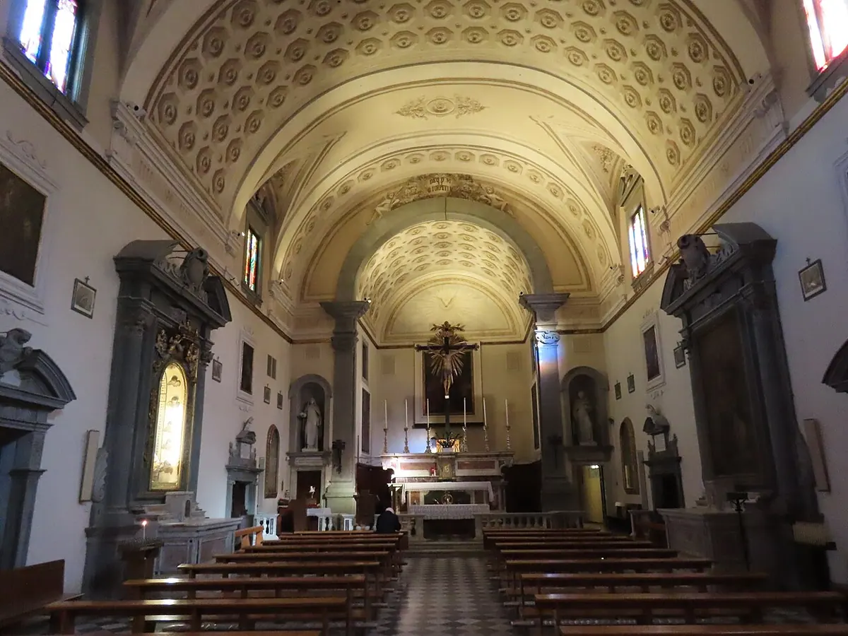 A white church nave with a coffered barrel vault, rows of wooden pews and side altars, leading to a high altar with a painting above it