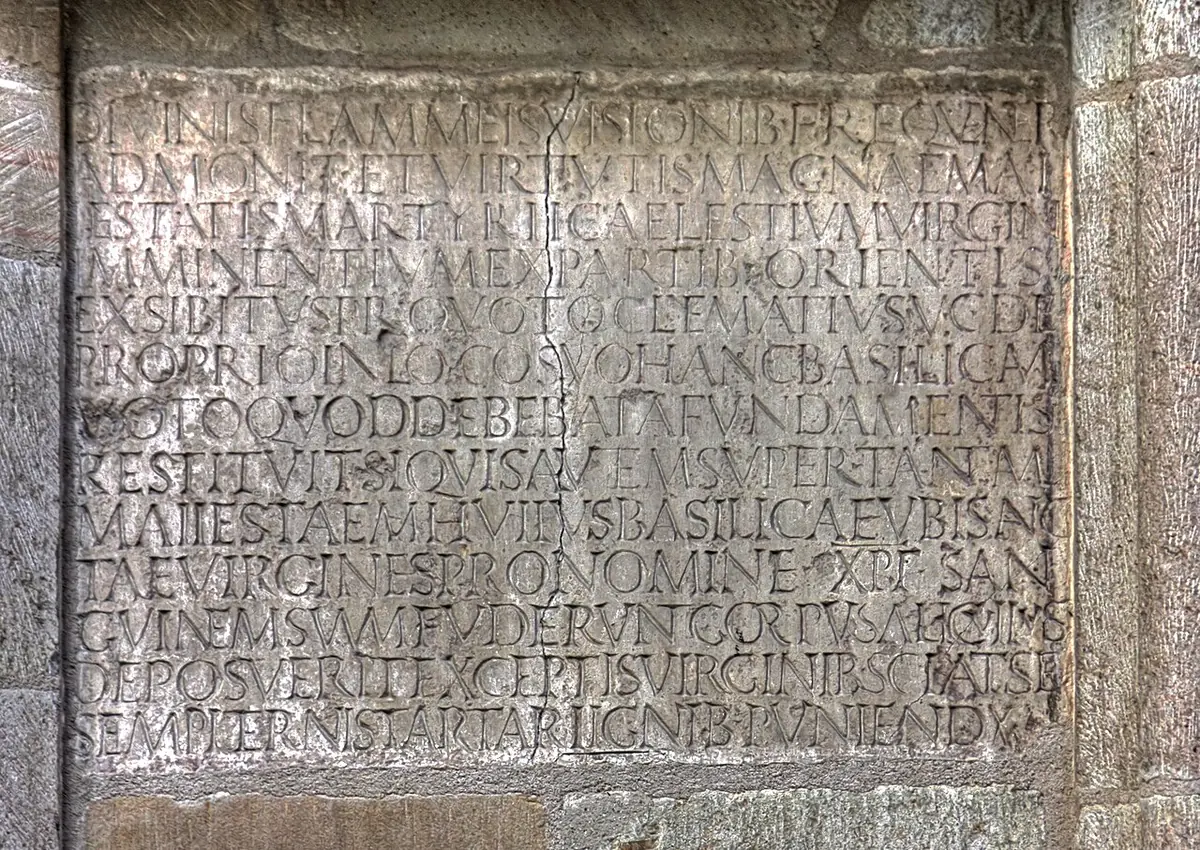 A worn gray stone slab set into a church wall, carved with fourteen lines of Roman capital letters