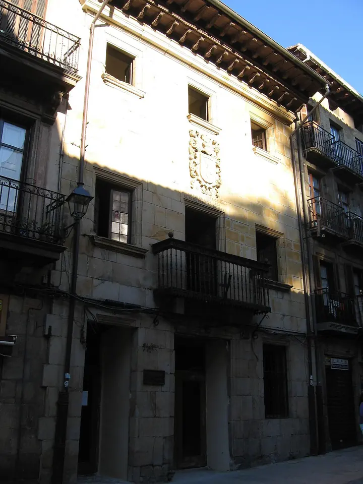 A tall stone town house with iron balconies, a carved coat of arms and wide wooden eaves on a narrow street in Elorrio