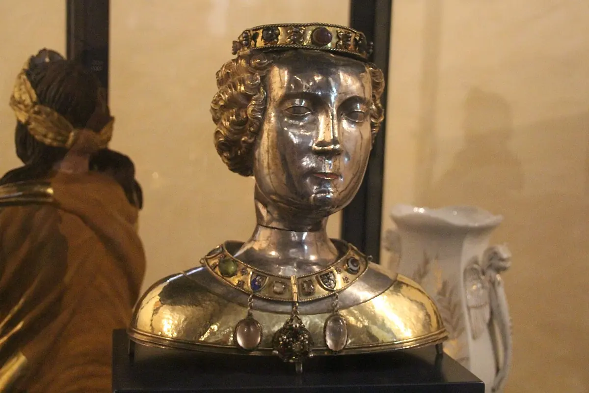 Silver reliquary bust of a young woman with gilded wavy hair and a jeweled crown, wearing a gilded jeweled collar, in a glass case
