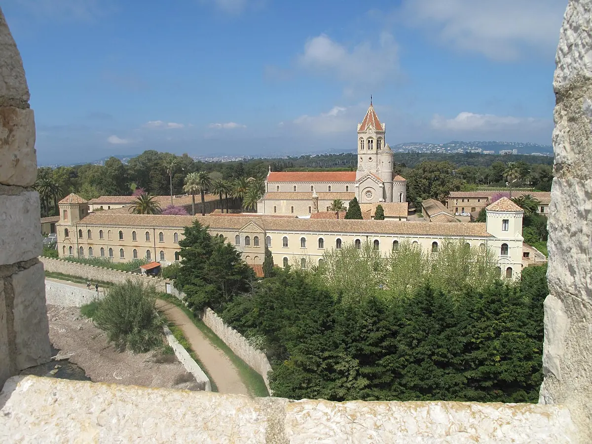 The cream-colored buildings and bell tower of Lérins Abbey among pines and gardens, seen between two stone battlements, with the sea and the coast of Cannes beyond