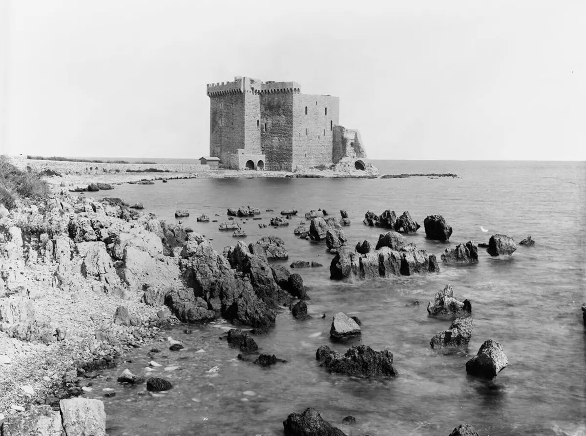 Old black-and-white photograph of the square stone tower of the fortified monastery of Lérins standing at the water's edge, with rocks scattered in the sea in front