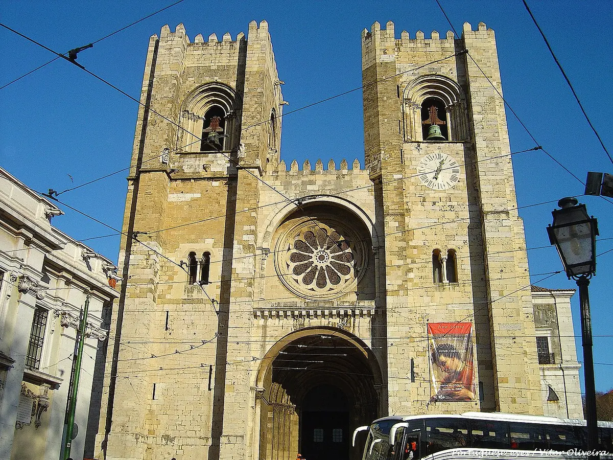 The fortress-like Romanesque facade of Lisbon Cathedral with two crenellated towers and a rose window, under a blue sky