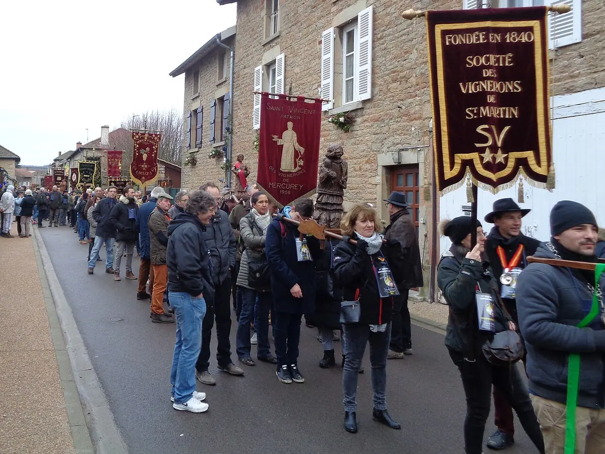 Winegrowers' societies march down a village street in winter carrying embroidered banners and a statue of St. Vincent during the Saint-Vincent Tournante festival