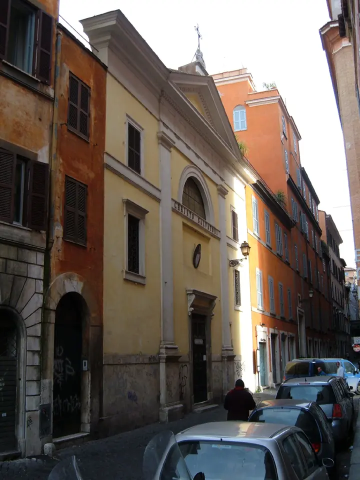 A narrow Roman street with the pale yellow facade of a small church squeezed between tall ocher and orange buildings