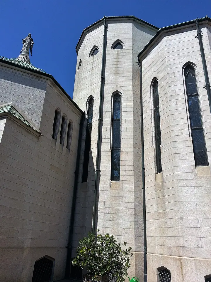 Tall white stone walls of a church apse with narrow pointed windows, seen from below against a deep blue sky, a statue on the roof