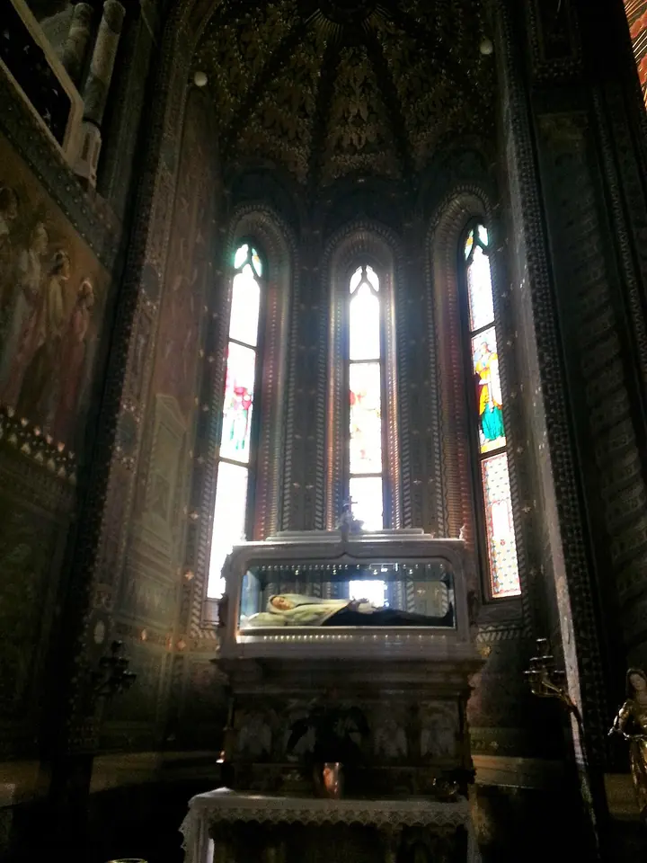 Glass case on a carved altar in a dark Gothic-style chapel lit by three tall stained-glass windows, the body of a sister in a black habit resting inside