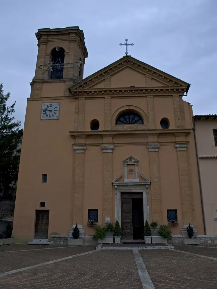 A pale ochre church with a classical pediment, pilasters, a round window and a square bell tower with a clock, behind a paved square