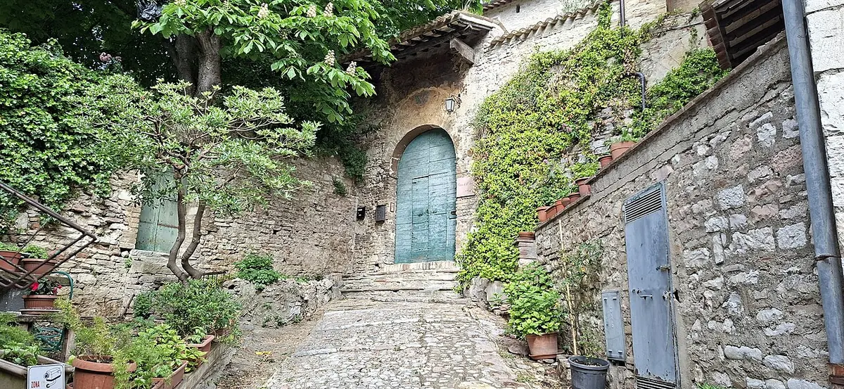 A cobbled lane climbing to a small stone church with a green wooden door under a little tiled porch, with ivy and potted plants on the old stone walls