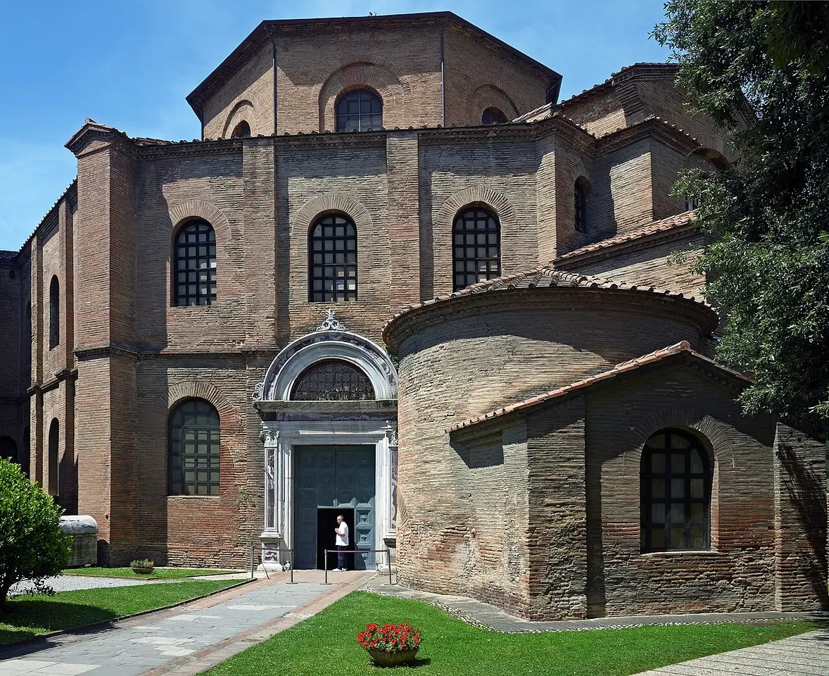 The octagonal brick Basilica of San Vitale with its stepped roofs and arched windows, seen from a green lawn on a sunny day