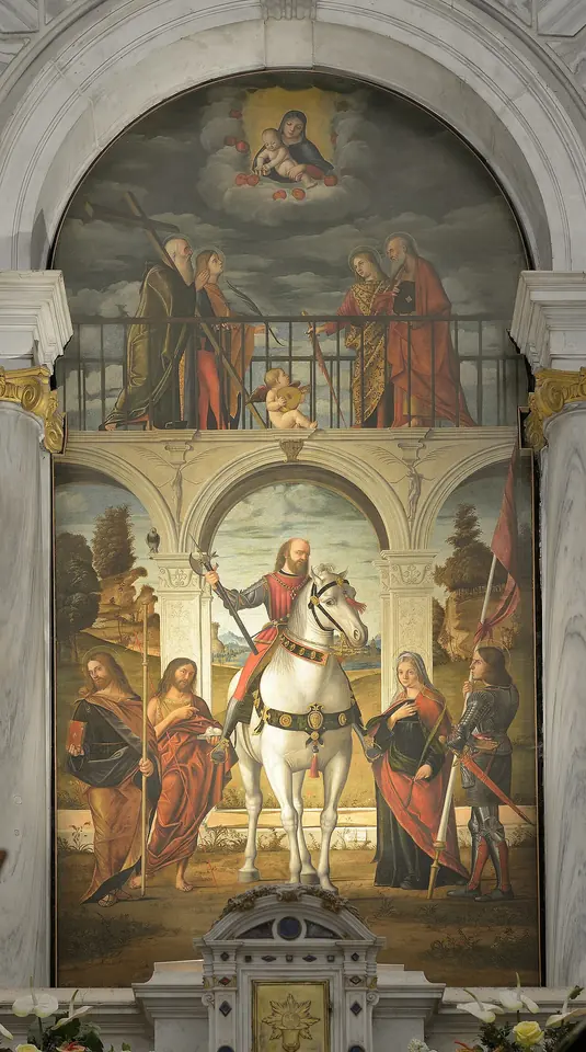 Tall Renaissance altarpiece of a knight in armor on a white horse in an arcaded loggia, surrounded by standing saints, with the Virgin and Child in the clouds above and more saints on a balcony