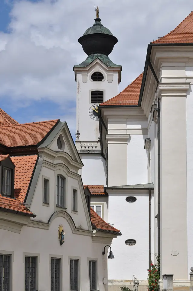 The white bell tower of a Baroque church with a green onion dome, behind the red-roofed buildings of a Bavarian abbey